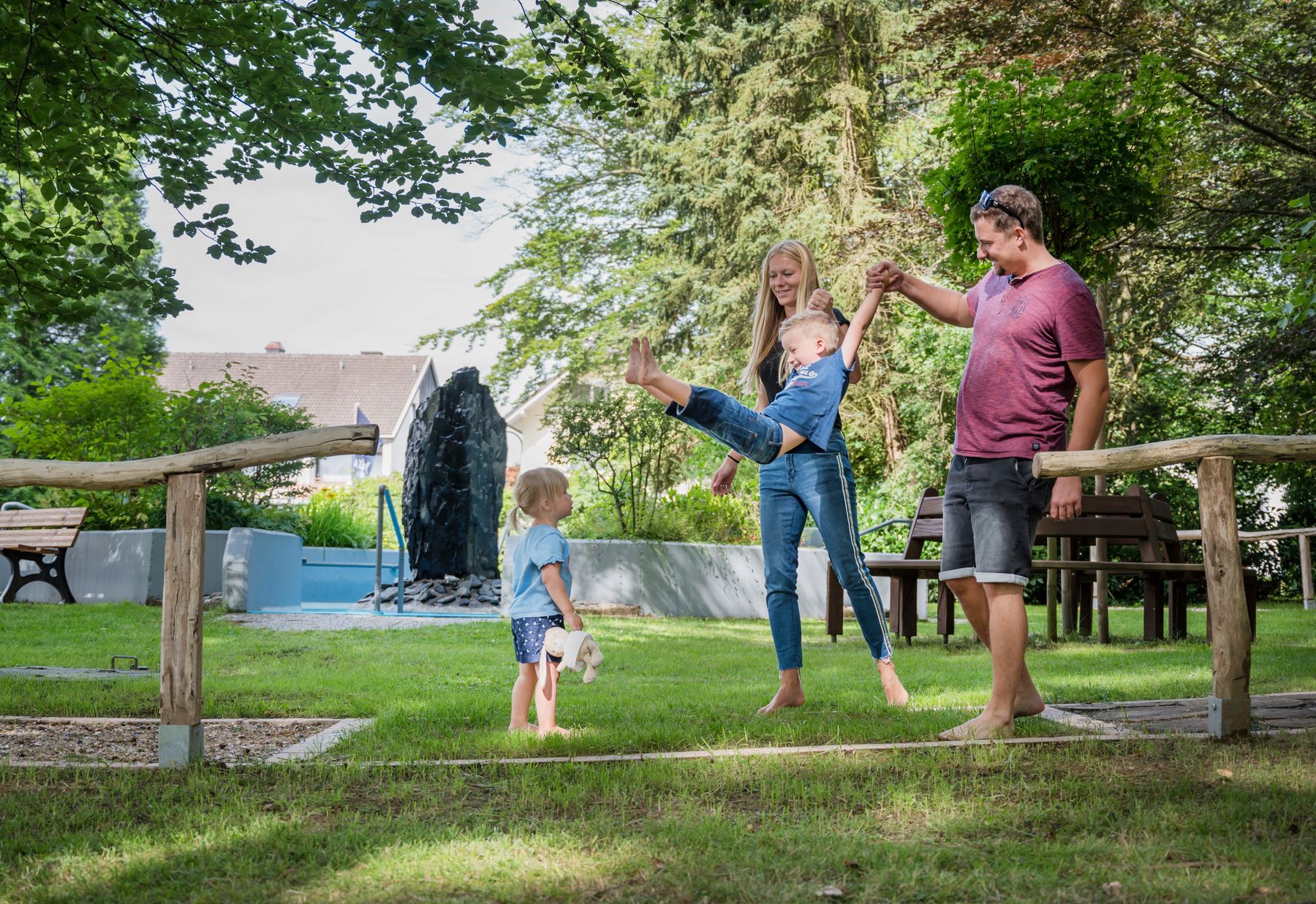 Eine glückliche vierköpfige Familie spielt in einem sonnigen Park. Die Eltern schwingen ein Kind an den Armen, während das andere Kind in der Nähe steht und ein Stofftier hält. Im Hintergrund sind Bäume und ein Wasserspiel zu sehen.